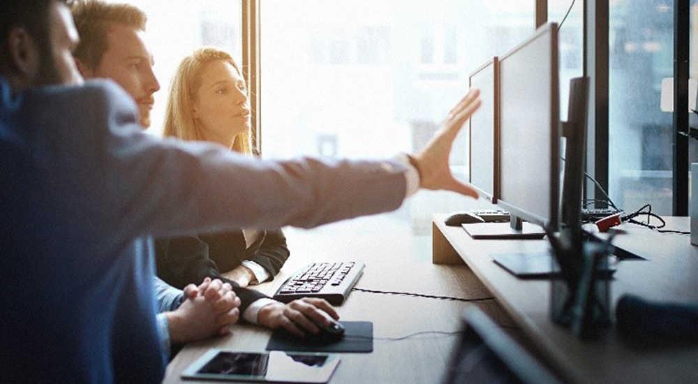 Business professional pointing to computer screen, talking to two others